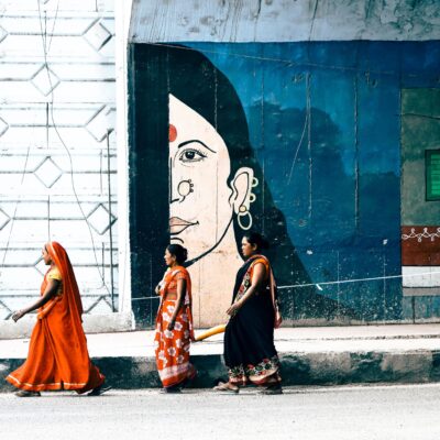 Two women in Indian walking in front of a large mural depicting half a woman’s face.